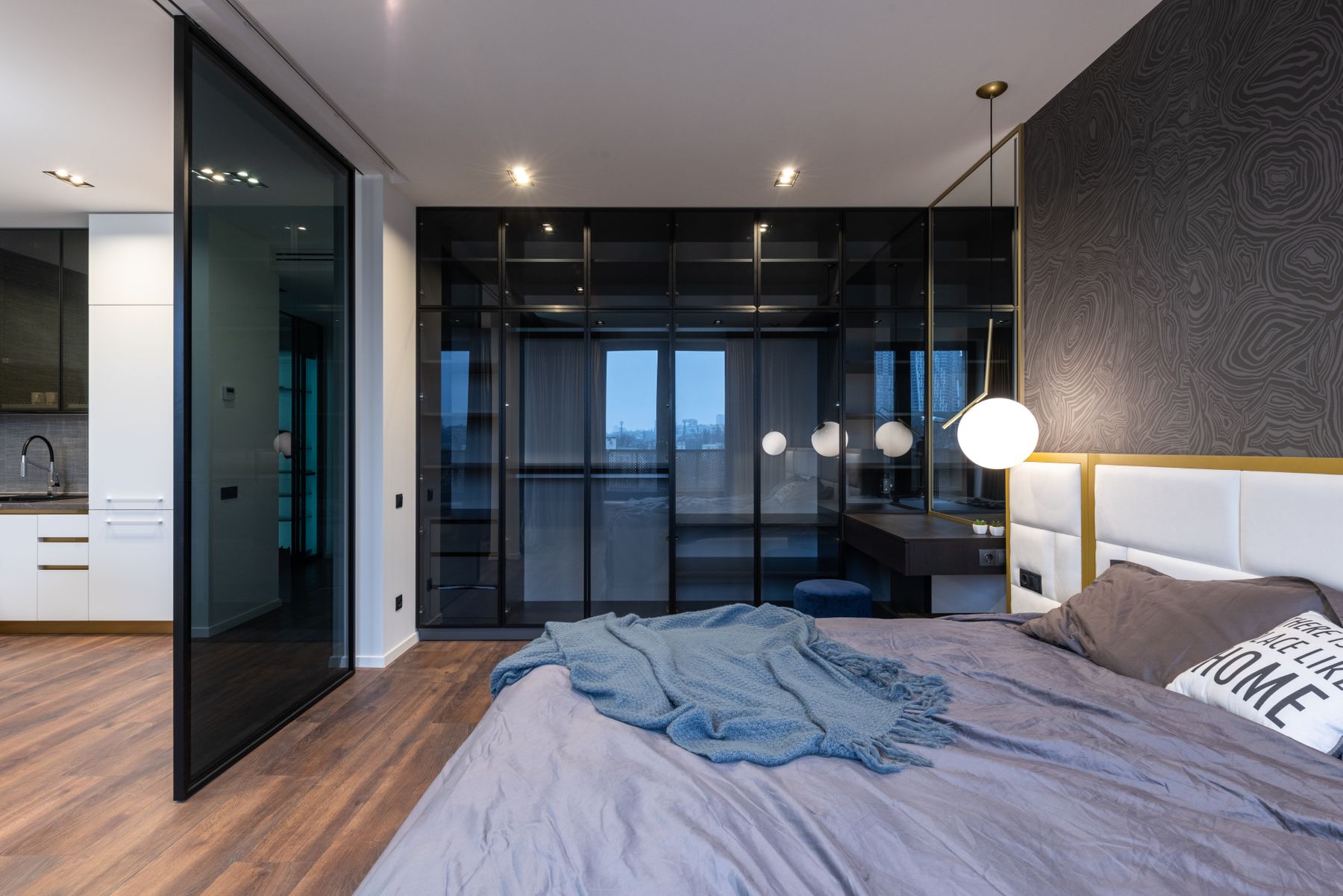 Schlafbereich mit grauer Bettwäsche, blauer Decke, Pendelleuchte und dunklen Glastüren