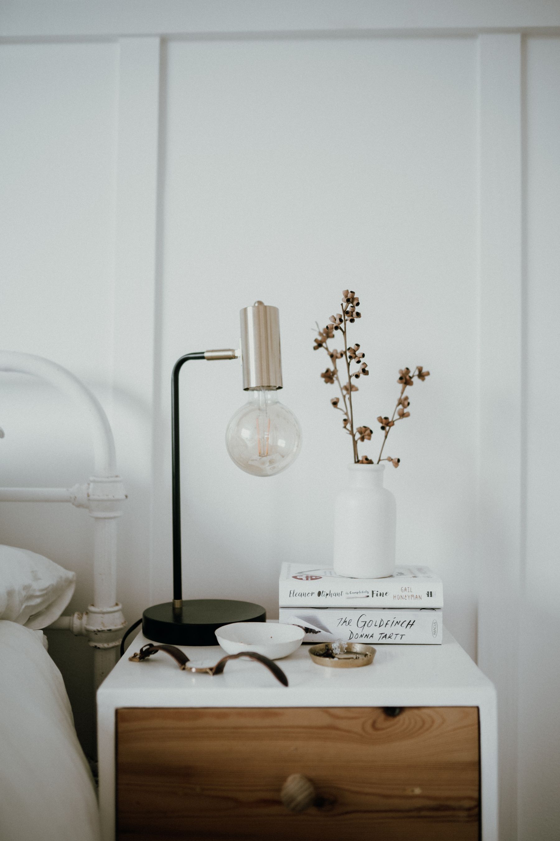 Nachttisch mit schlichter Leuchte, weißer Keramikvase, Zweigen und Büchern